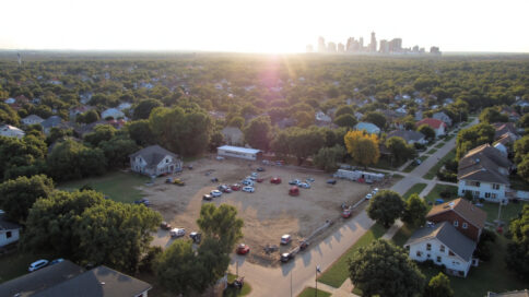 Aerial view of a Texas neighborhood highlighting diverse property types with a construction site in the foreground and a city skyline in the background under warm sunlight.