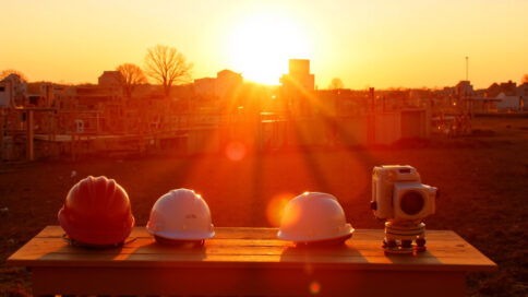 A construction site at dawn showcasing a Texas landscape with architectural blueprints on a wooden table, hard hats, and surveying equipment nearby, illuminated by golden sunlight casting long shadows.