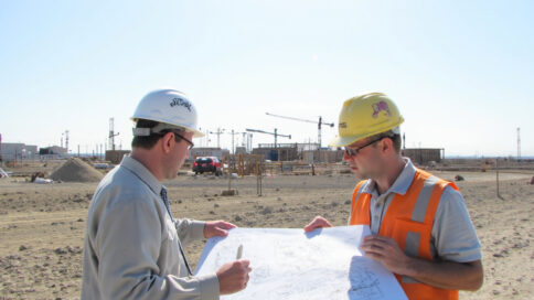 A construction site in Texas with cranes and buildings, a site manager reviewing plans under harsh sunlight with a cellular tower in the background.