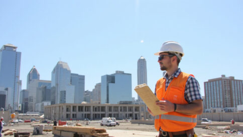 Worker inspecting building materials at a construction site in Texas with skyscrapers in the background and safety equipment visible.