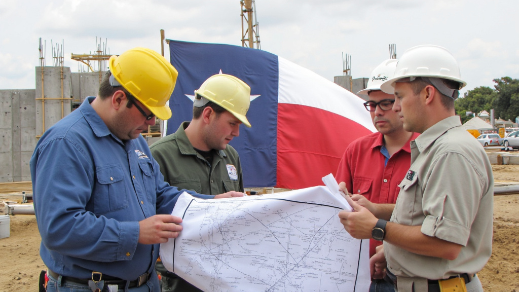Official inspectors in hard hats examining blueprints at a construction site with the Texas state flag visible.