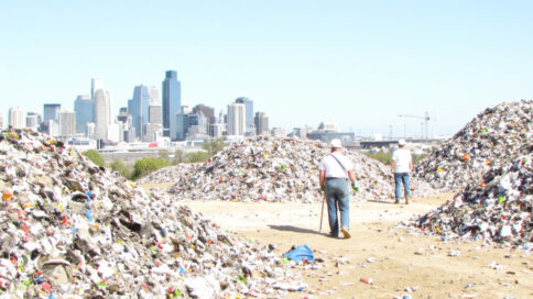 Construction site with recycled materials, workers in hard hats, and Texas skyline in background under bright sunlight.