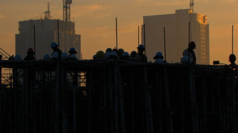 Construction site in Texas with empty hard hats on scaffolding and a lone worker, set against an unfinished skyscraper during a golden sunset.