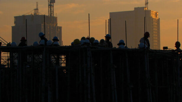 Construction site in Texas with empty hard hats on scaffolding and a lone worker, set against an unfinished skyscraper during a golden sunset.
