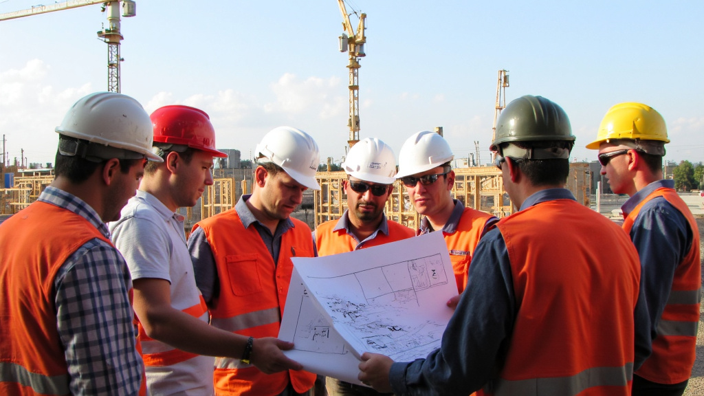 Construction Professionals Discussing Blueprints Diverse group of construction professionals in hard hats and safety vests discussing blueprints on a construction site with cranes and scaffolding in the background.