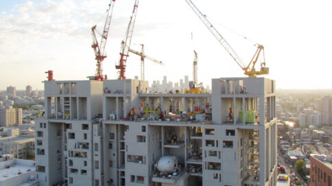 Modern construction site with prefabricated building components, workers assembling modular sections, cranes lifting factory-made units, sunlit urban backdrop.