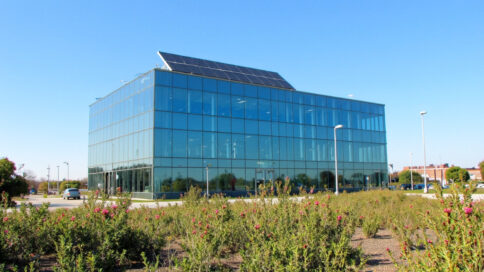 Modern green office building in Texas with a glass facade reflecting the blue sky and visible solar panels, surrounded by native landscaping.