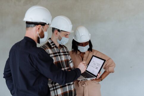 Three construction workers discussing building plans on a laptop at a site.