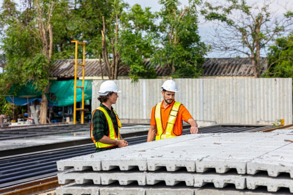 Two construction workers in hard hats discuss at a building site outdoors.
