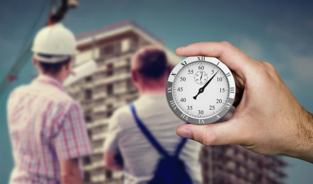 Hand holding stopwatch with blurred construction workers and building background.