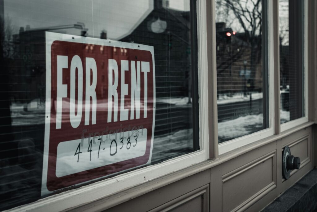 "A 'For Rent' sign is displayed in a shop window on a city street."