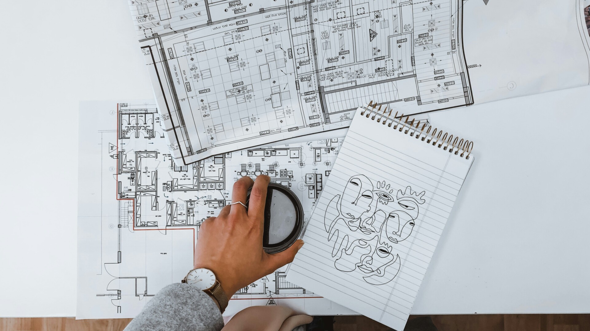 Creative Architectural Workspace A hand holding a cup above architectural plans and a notepad with abstract sketches.