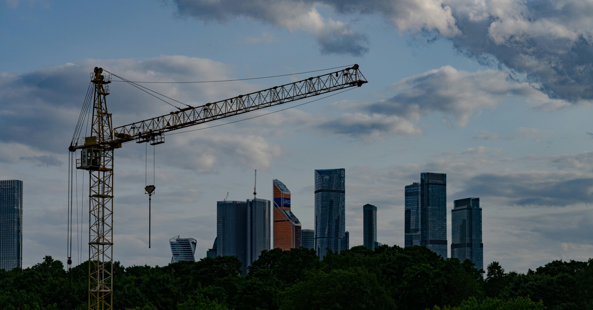 Construction Crane and Urban Skyline A construction crane overlooks a city skyline with trees in the foreground and clouds in the sky.