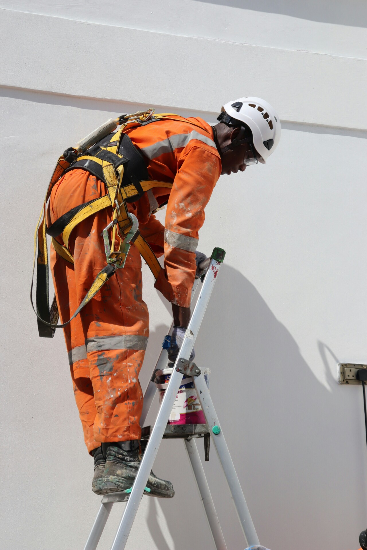 Safety First: Construction Worker at Height A construction worker in an orange uniform and safety gear using a ladder to paint a wall.