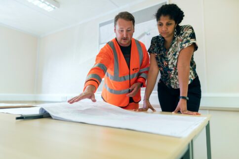 Two professionals discussing plans at a table, one wearing a safety vest