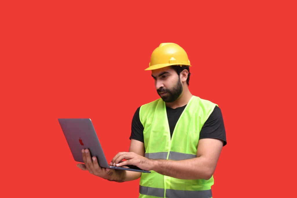 A construction worker in a yellow safety helmet and green reflective vest using a laptop.
