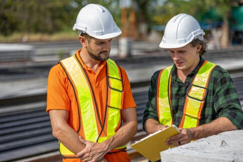 Two construction workers in safety gear discussing plans at a building site.