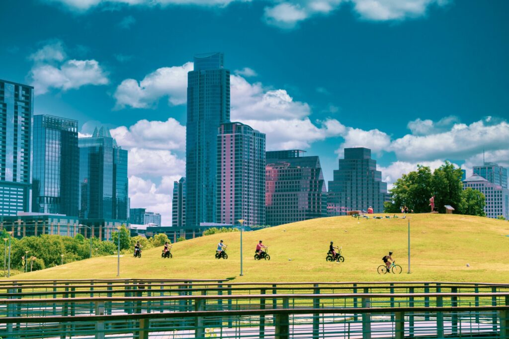 A group of cyclists rides across a grassy hill in an urban park.