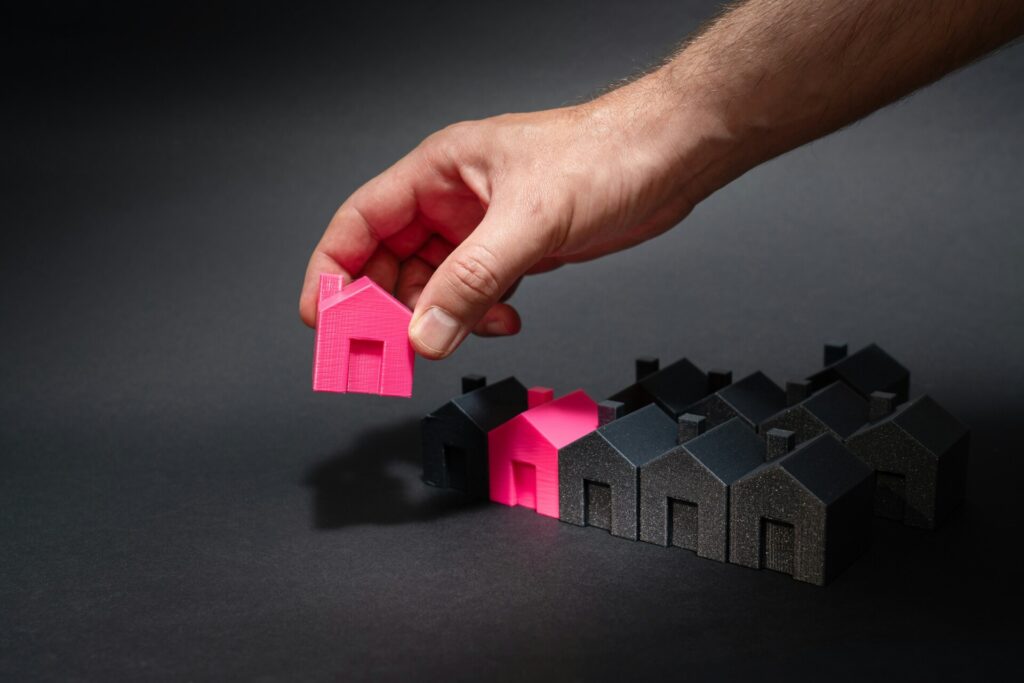 A hand places a bright pink house model among black house models.