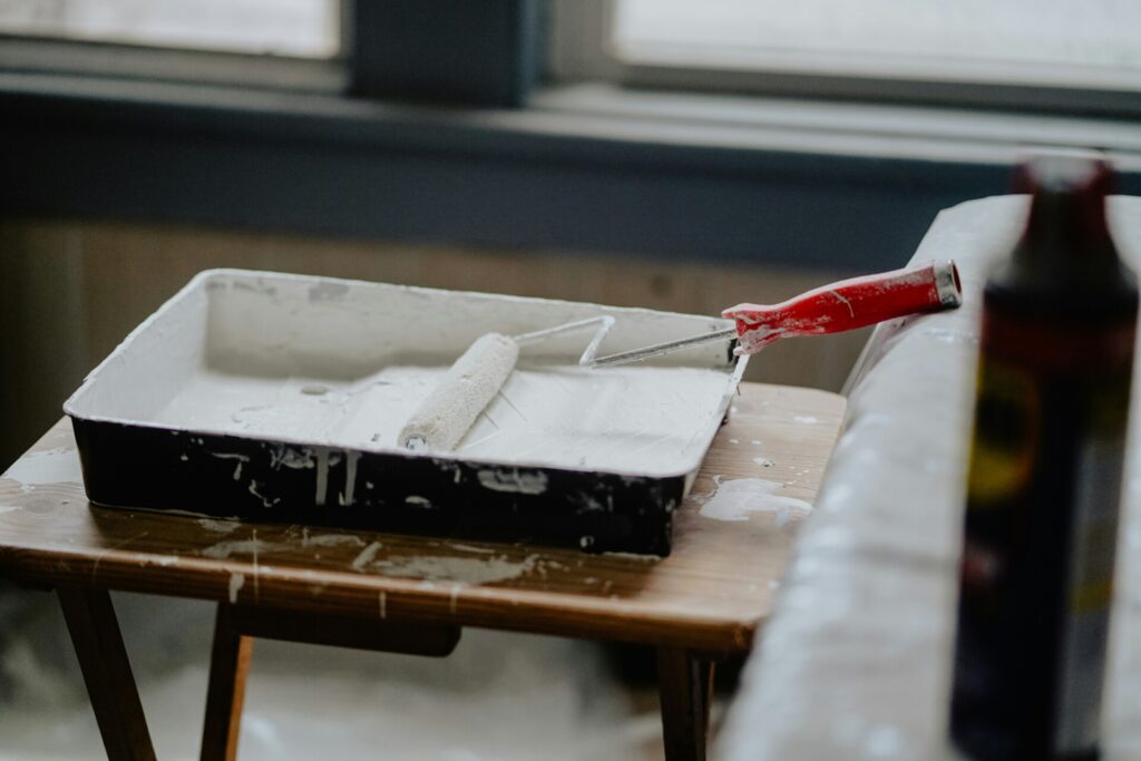 Paint roller and tray with white paint on a wooden stool by a window.