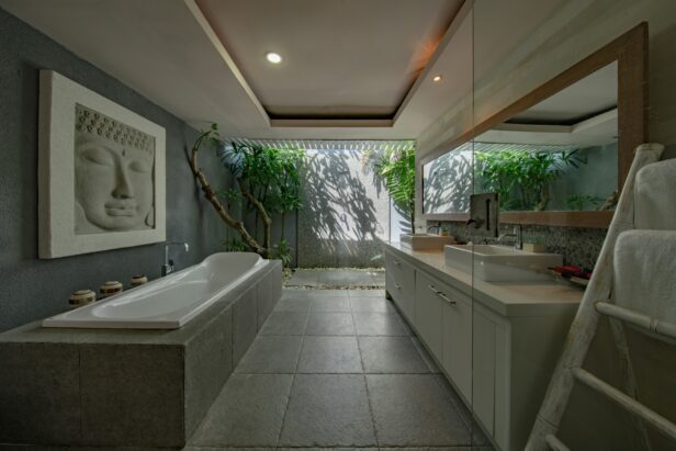 A serene and luxurious bathroom featuring a large bathtub, a stone wall art of a Buddha face, and lush greenery visible through a window.