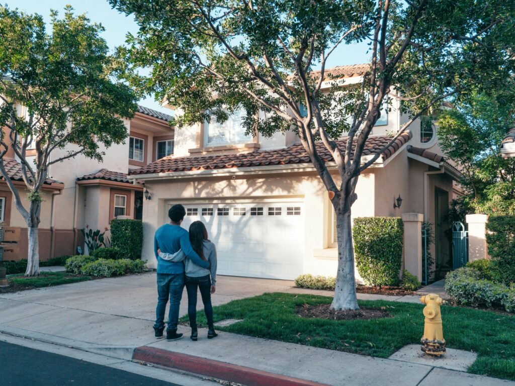 A couple stands together looking at a suburban house on a sunny day.
