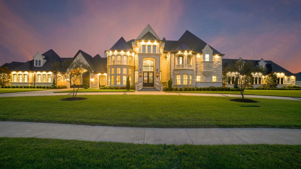 A grand mansion illuminated at dusk with a vast green lawn in front.