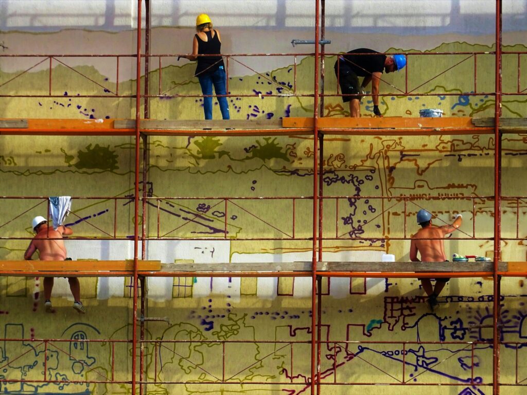Four workers paint a mural on a large wall using scaffolding.