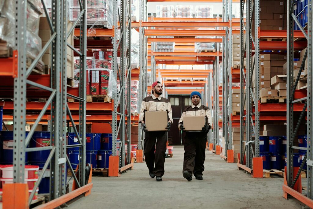 Two workers carrying boxes in a large warehouse with metal shelves.