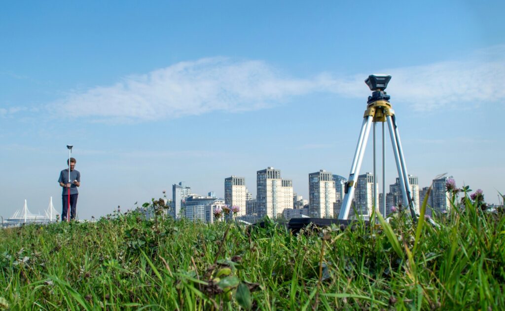 A surveyor uses equipment in a grassy field with a cityscape behind.