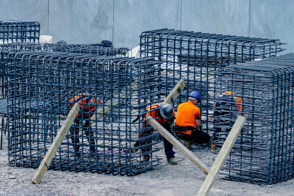 Workers in orange vests assemble large metal rebar structures at a construction site.
