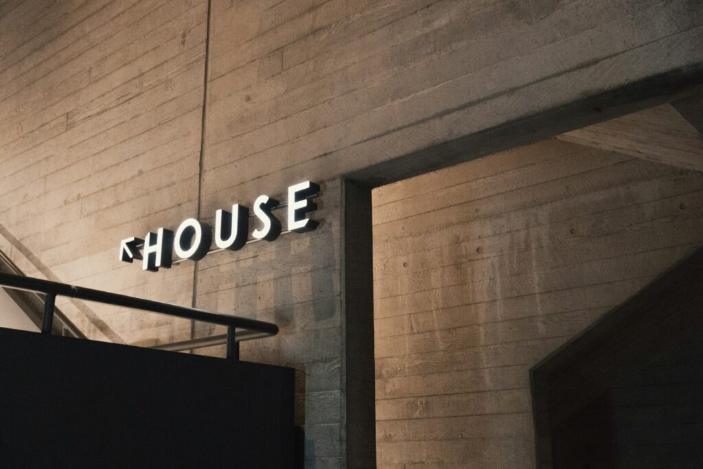 Illuminated "HOUSE" sign on a textured concrete wall in a dim setting.