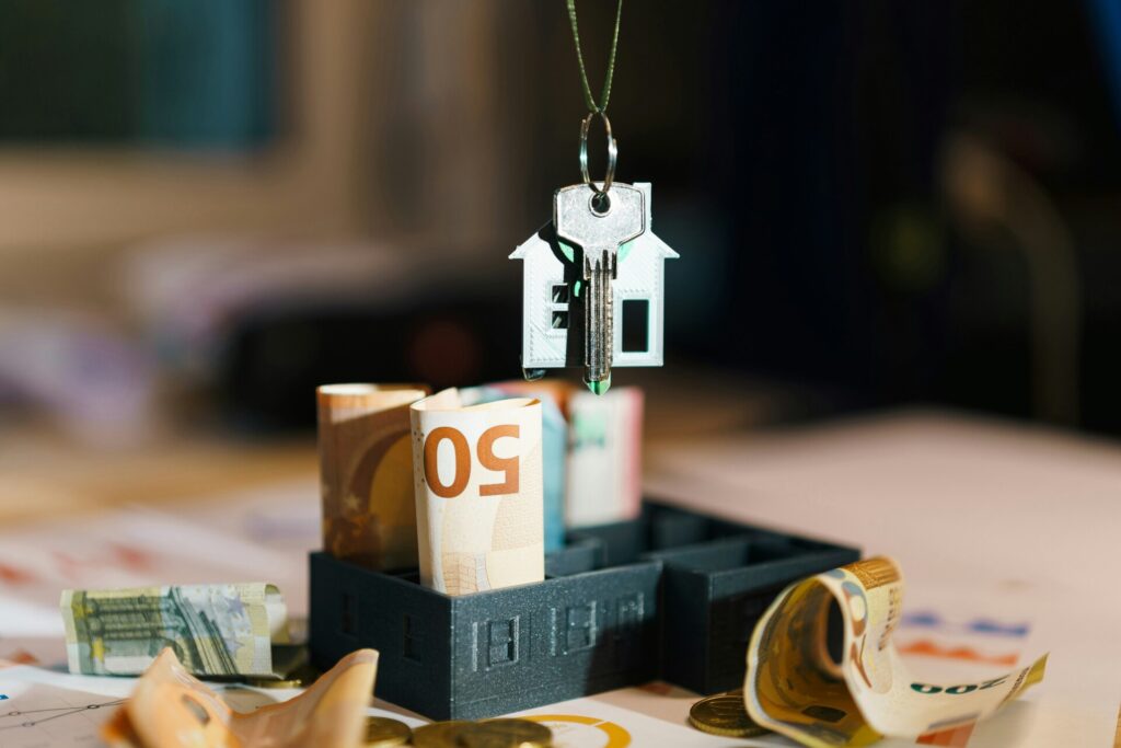 A house key fob hovers over rolled banknotes in a miniature house model.