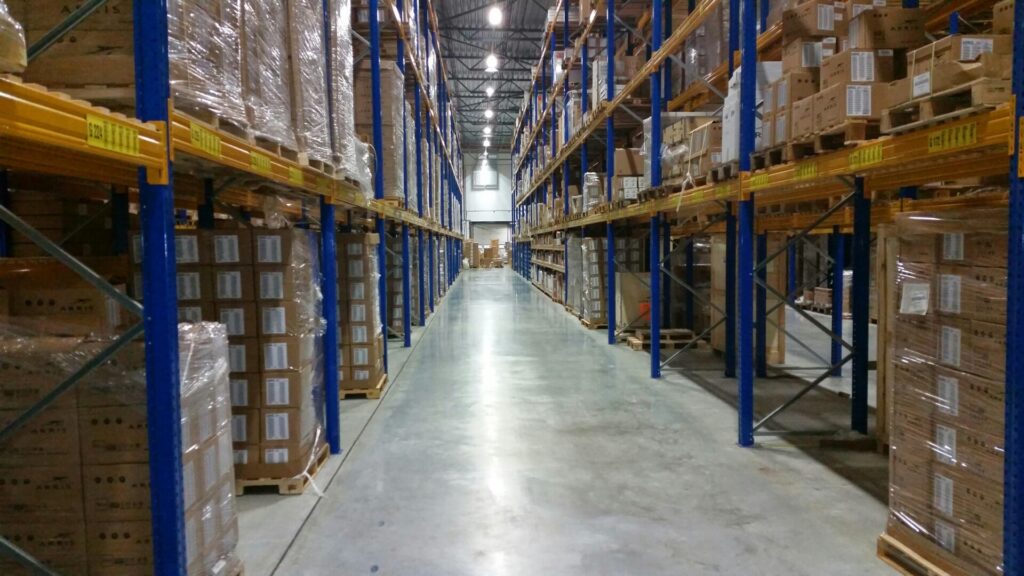 Warehouse aisle with stacked boxes on tall shelves under bright lighting.