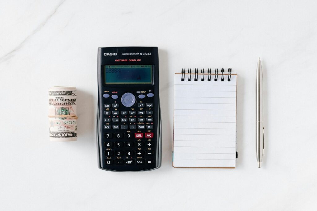 A calculator, rolled banknotes, notepad, and pen on a white surface.