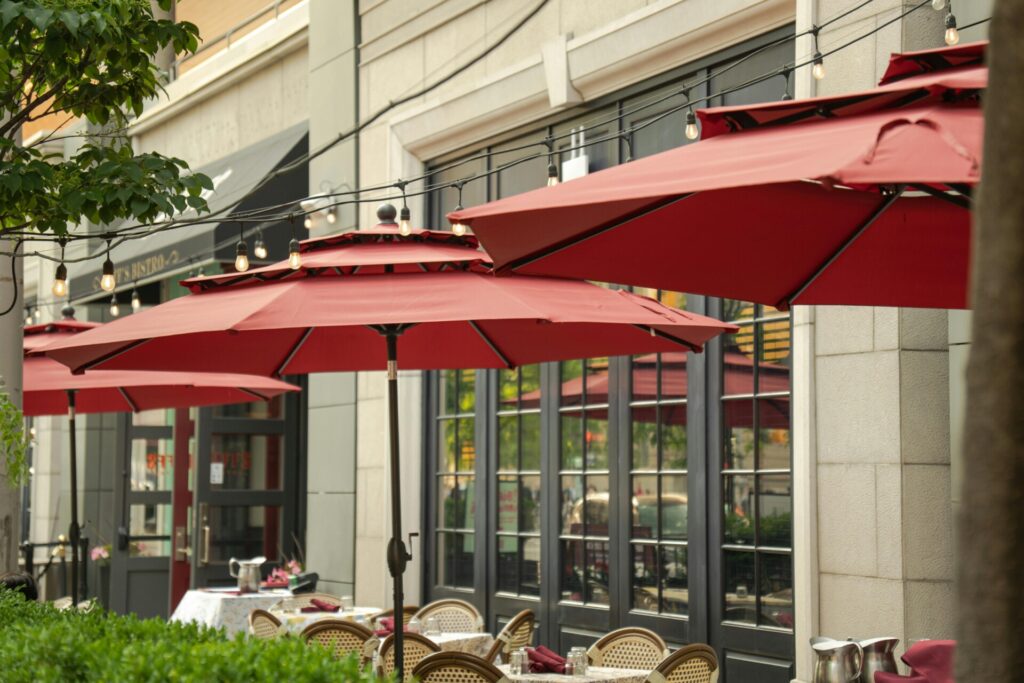 Outdoor cafe with red umbrellas and string lights above tables.