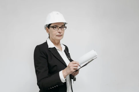Woman in a hard hat with a clipboard stands against a blank background.
