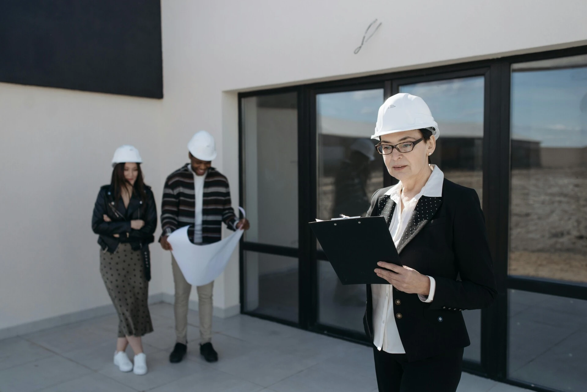 Three construction professionals wearing hard hats review plans outside a building.