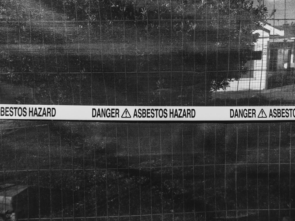 Black and white image of a fence with "DANGER ASBESTOS HAZARD" tape.