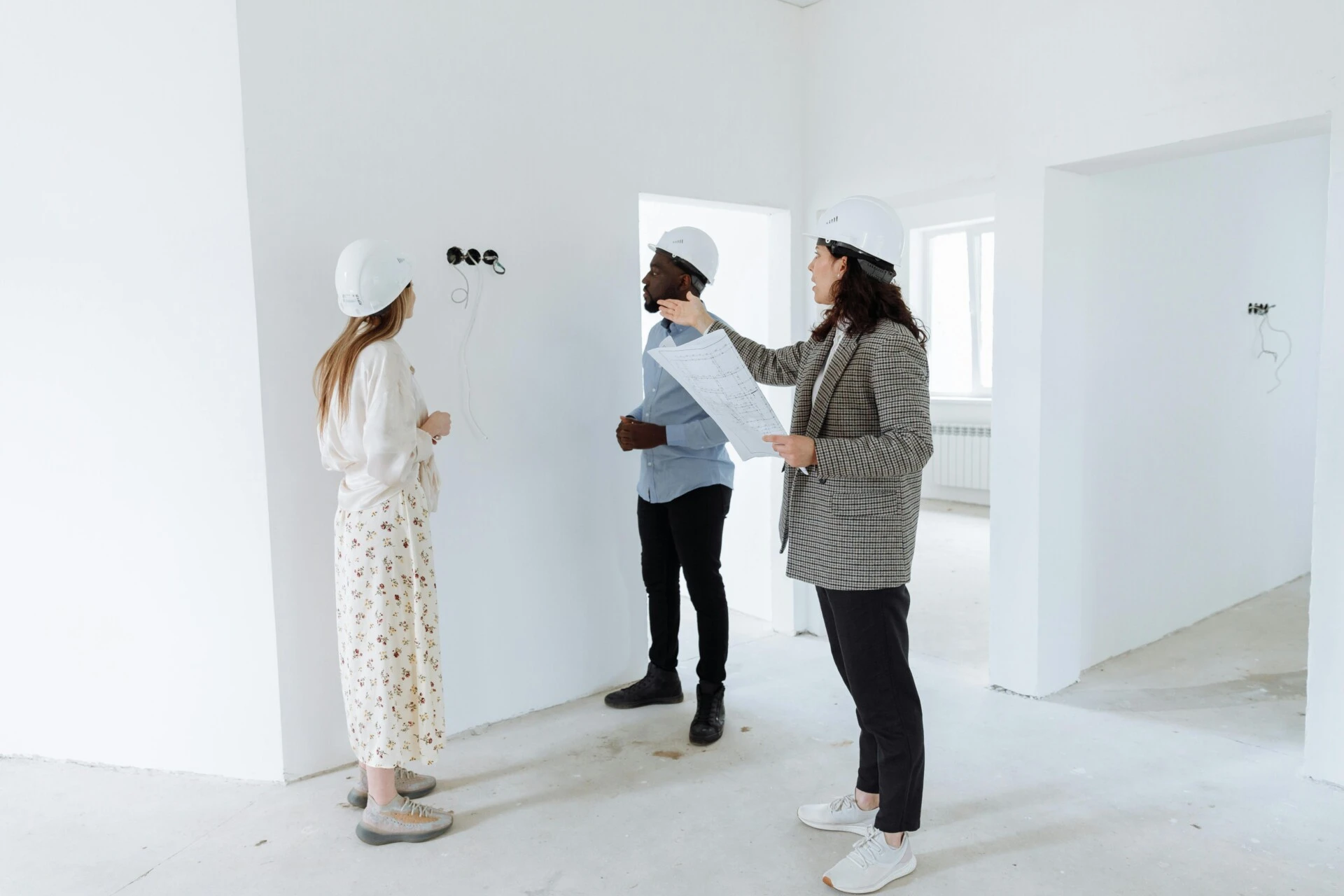 Three people in hard hats discussing electrical wiring in a white room.