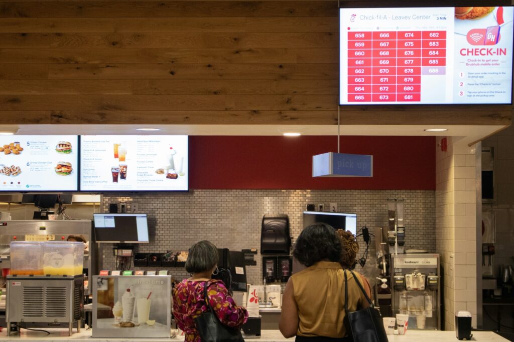 Three customers stand at a fast-food counter with digital menu displays.