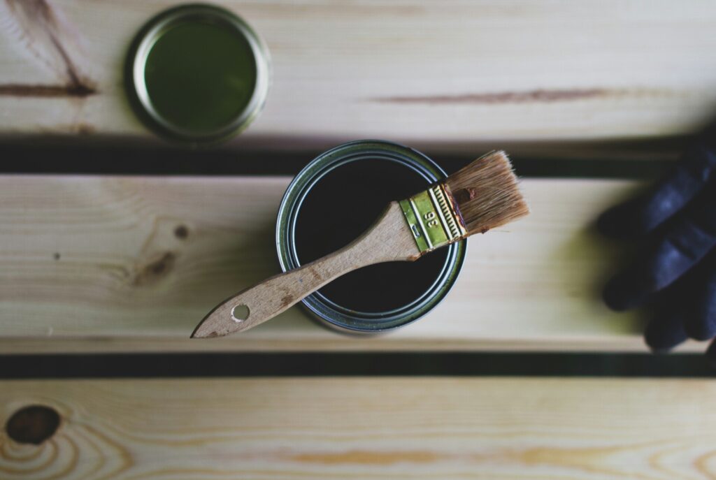 A paintbrush rests on an open can of paint on wooden boards.