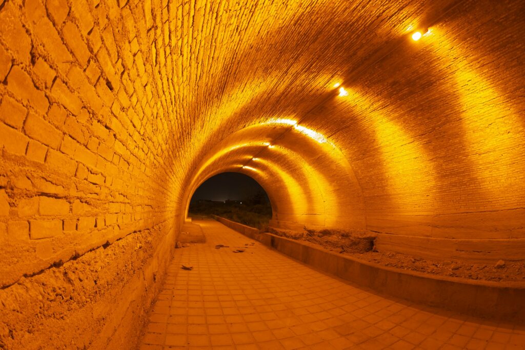 A warmly lit brick tunnel with a pathway leading to the outdoors.