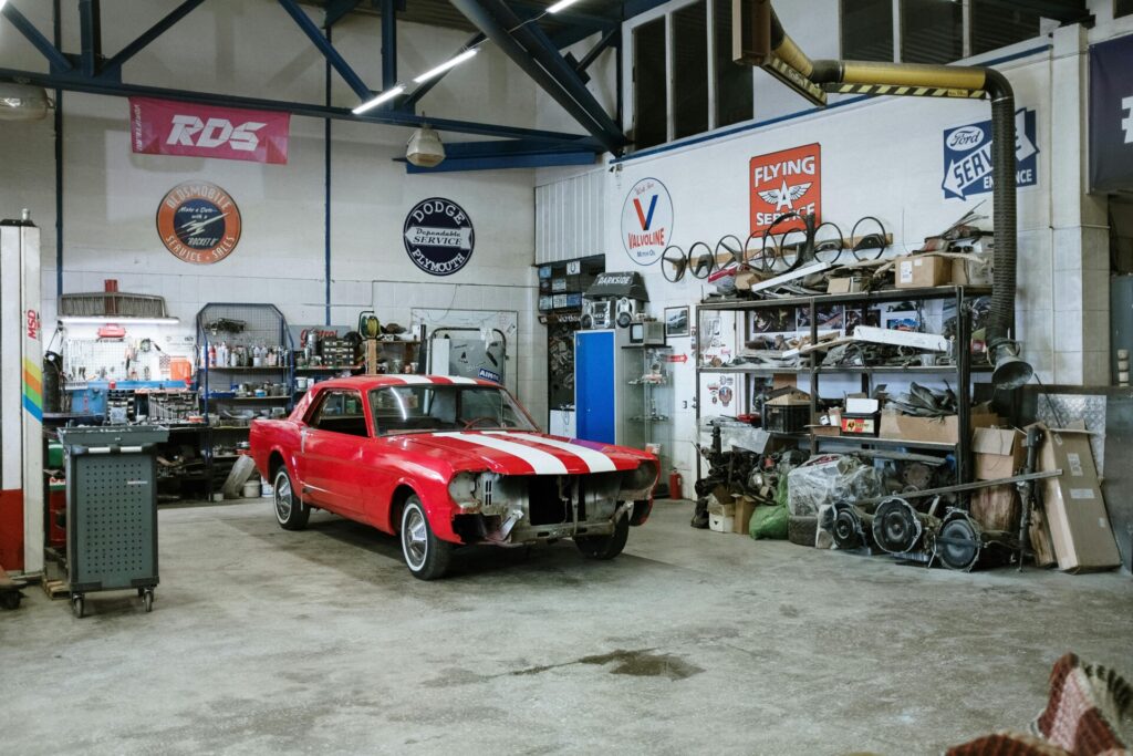 A red classic car under restoration in a cluttered auto garage.