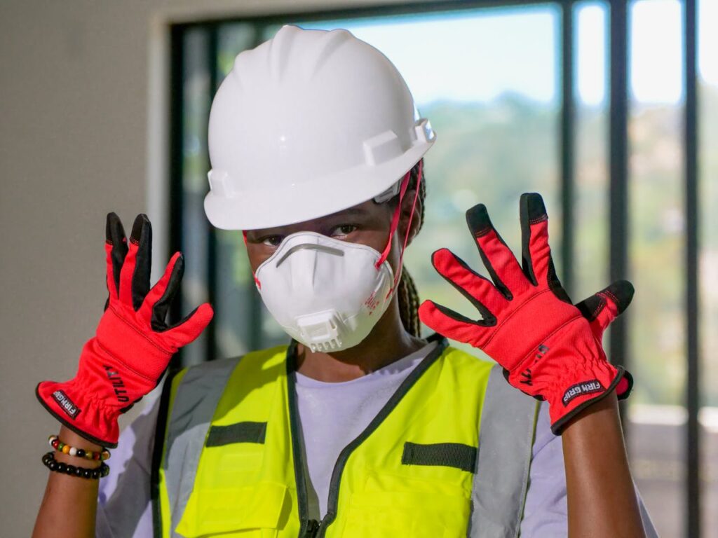 Construction worker with safety gear, including helmet, mask, and gloves, poses indoors.