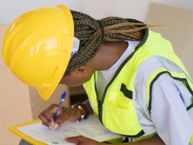 Person wearing a yellow hard hat and vest writing on a clipboard.