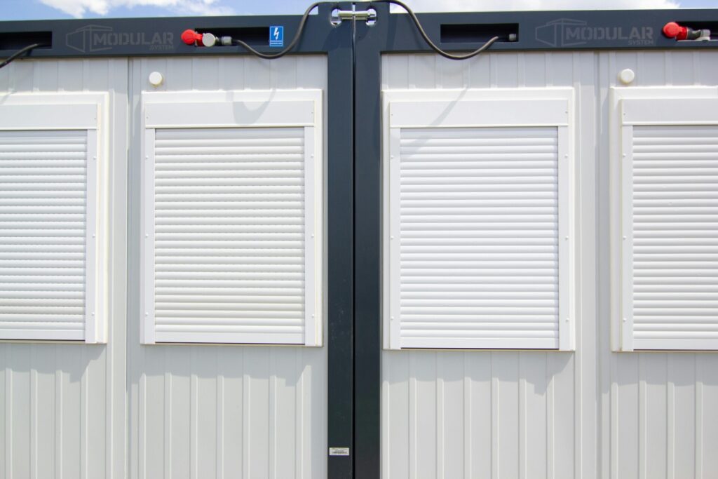 Modular building wall with closed white shutters and electrical connections on top.