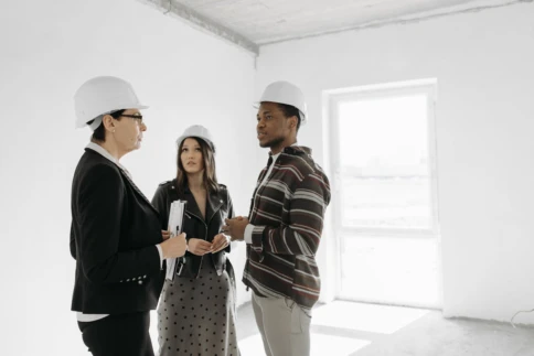 Three people wearing hard hats discuss plans inside a building under construction.