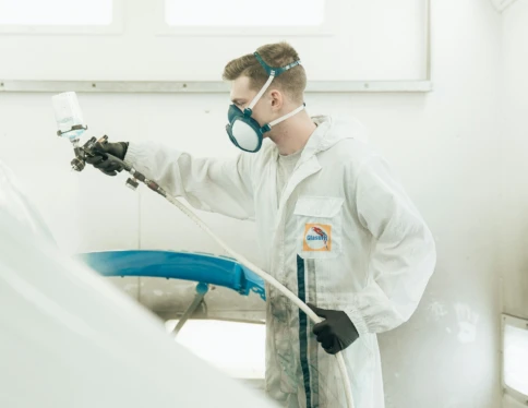 A person in protective gear spray-paints a car part in a workshop.