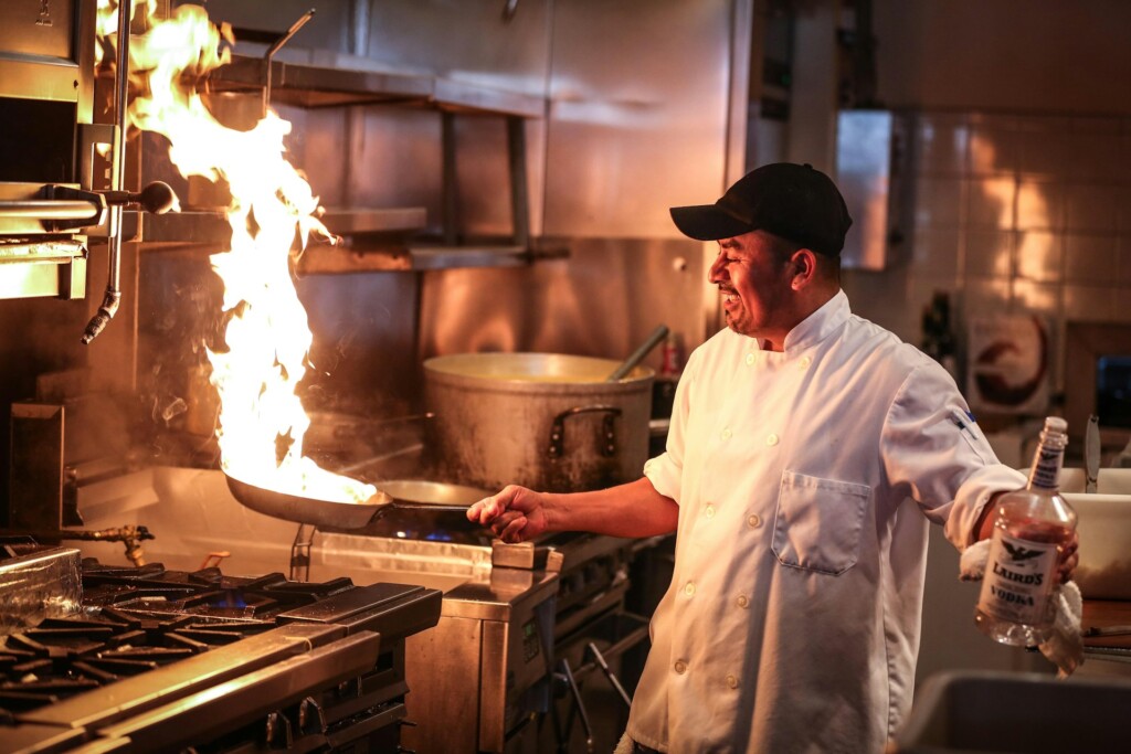A chef creates a dramatic flame in a kitchen with a frying pan.